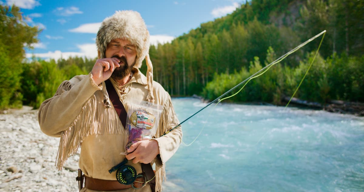 A man on a riverside shore holding a fishing pole and eating beef jerky with a forest mountain side in the background.